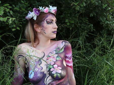 A woman painted with putple flowers against a green leafy backdrop.