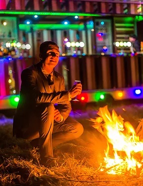 A smartly dressed man at a campfire, with a brightly lit bar behind him.